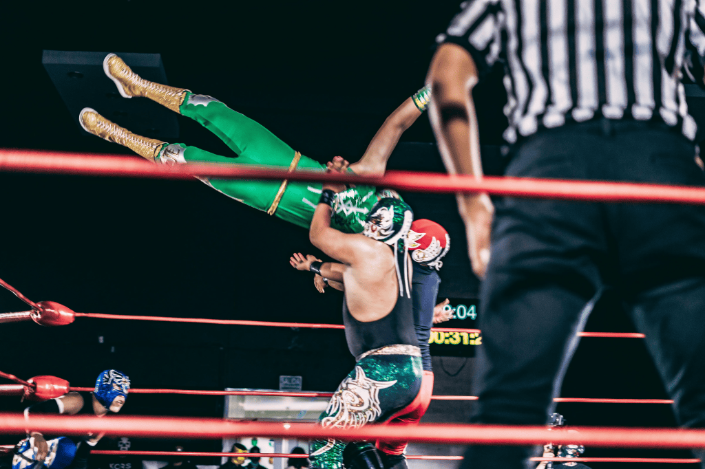 A wrestler in green and gold gear is being lifted off the ground by another wrestler in black and silver gear during a wrestling match. A referee stands in the foreground observing the action.